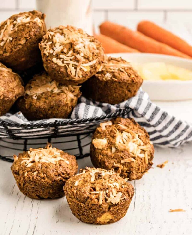 A basket lined with a striped cloth holds several morning glory muffins topped with shredded coconut. Three muffins sit in front on a white surface. In the background are whole carrots and a bowl of pineapple chunks.