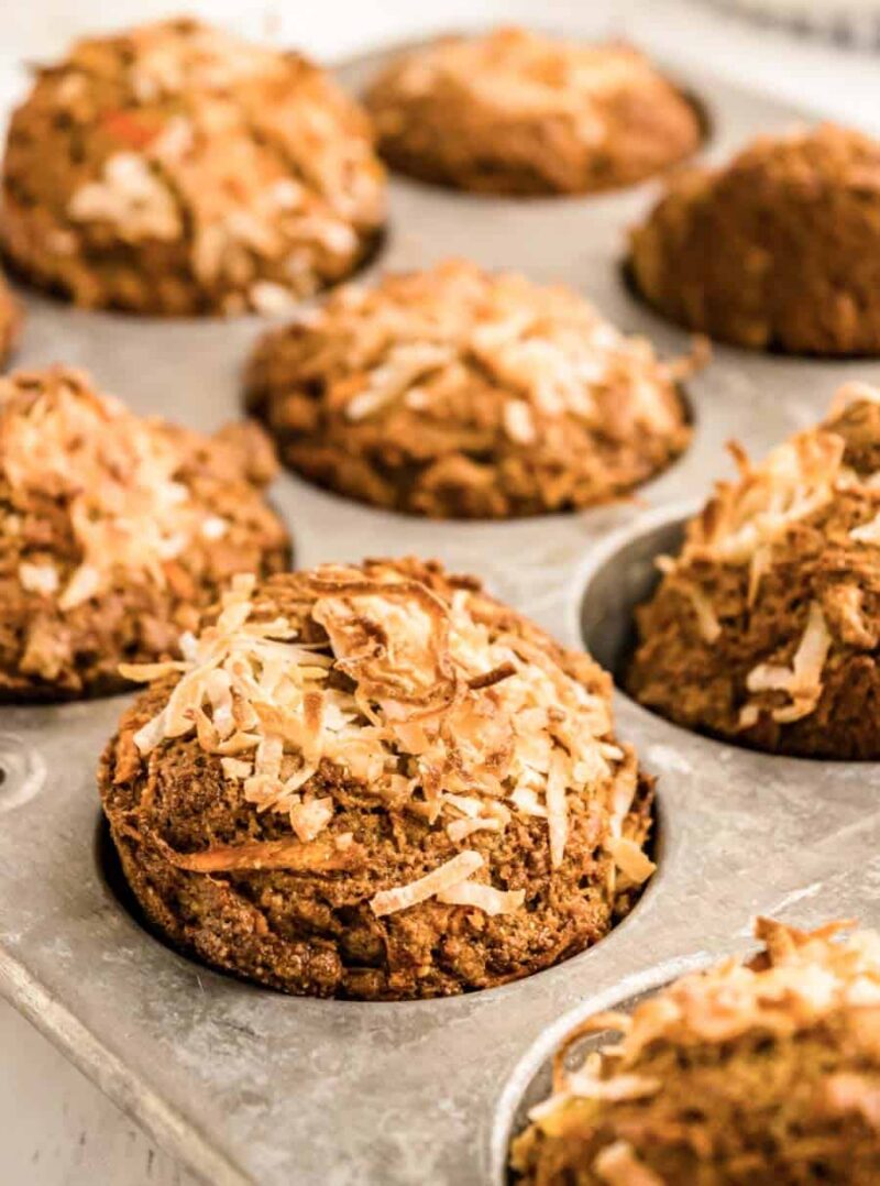 A close-up of freshly baked morning glory muffins topped with shredded coconut, still in a metal muffin tin. The golden brown muffins are textured, with a few in sharp focus and others softly blurred in the background.