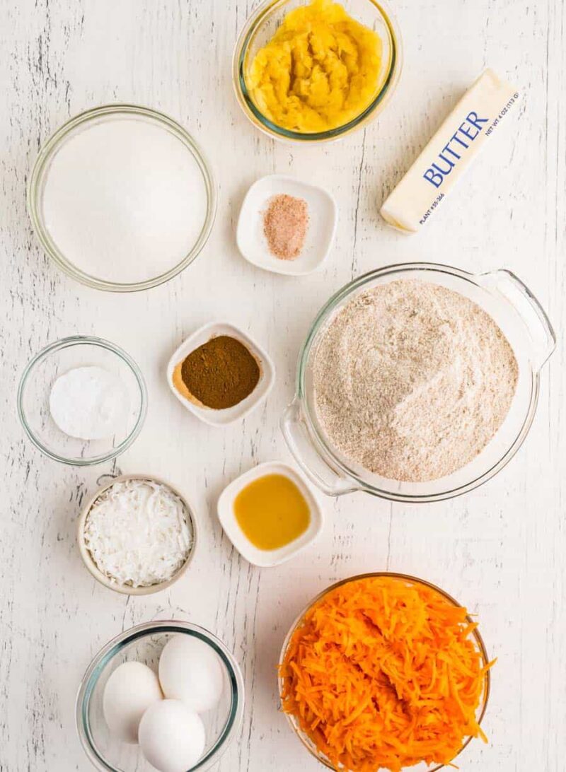 Carrot cake ingredients on a white surface, including grated carrots, eggs, flour, butter, sugar, shredded coconut, mashed fruit, baking powder, spices, and vanilla in small bowls—perfect for carrot cake or morning glory muffins.