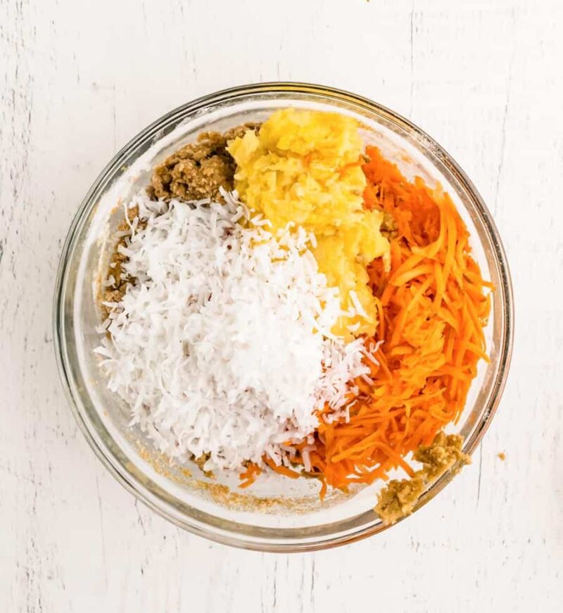 A glass bowl on a white surface containing the unmixed ingredients for morning glory muffins: shredded coconut, grated carrot, mashed banana, and brown sugar.