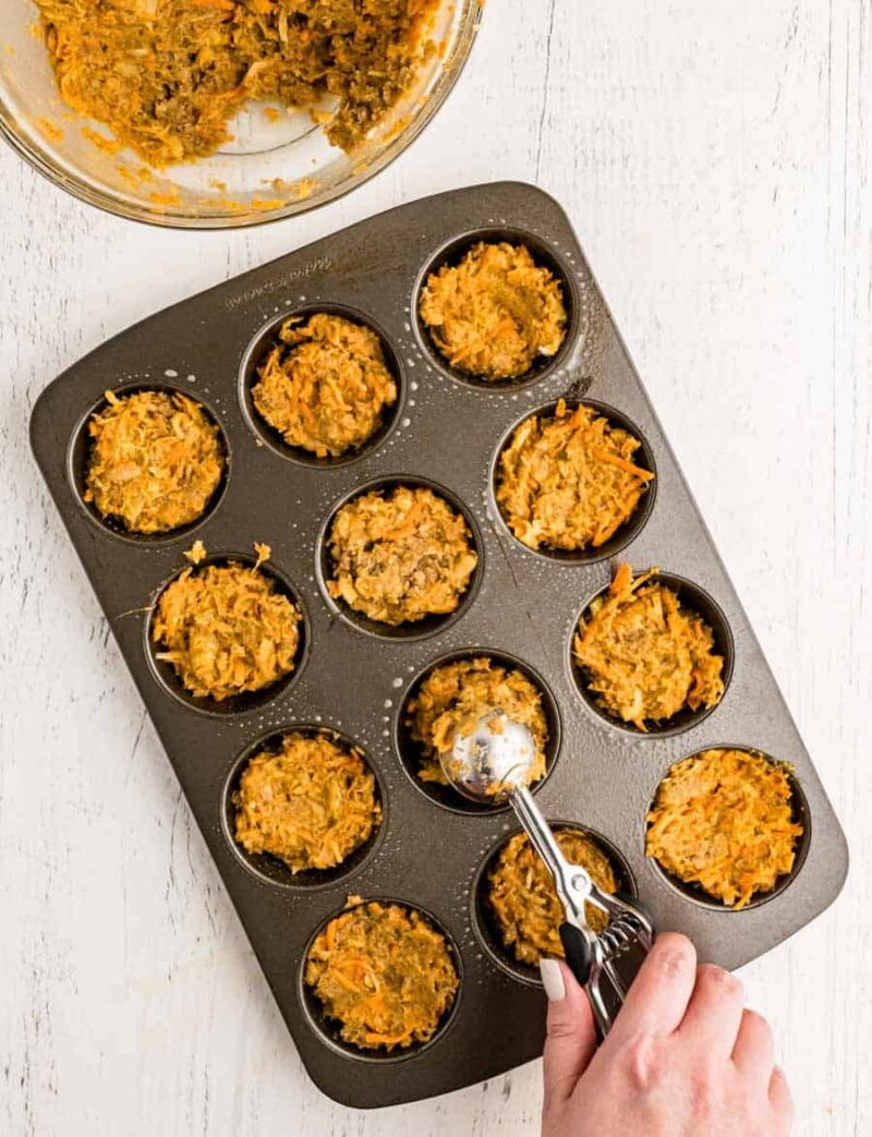 A hand uses a metal scoop to portion morning glory muffin batter—a carrot and oat mixture—into the cups of a nonstick muffin tin on a white surface. A glass bowl with leftover mixture sits in the top left corner.