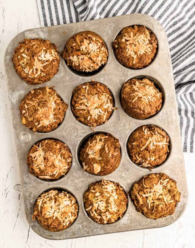 A metal muffin tin holds twelve baked morning glory muffins topped with shredded coconut. The tin sits on a white surface next to a gray and white striped cloth.