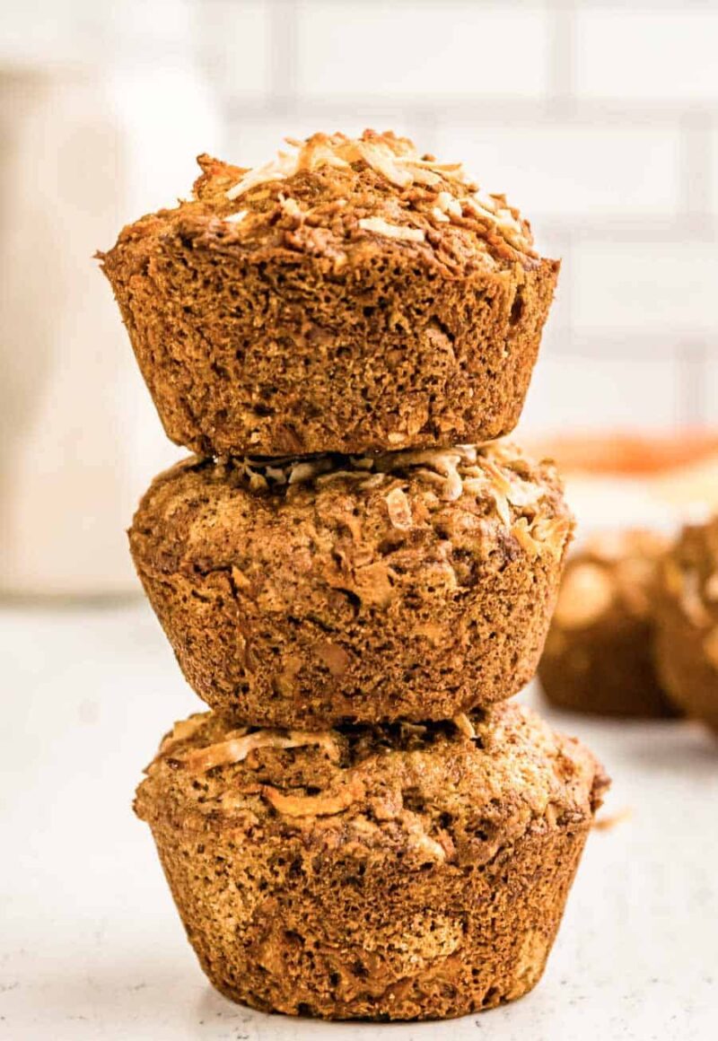 Three bran muffins, reminiscent of morning glory muffins, are stacked on a light surface. Behind them, a blurred muffin and a white background set the scene. The golden brown muffins feature a textured, crumbly top.