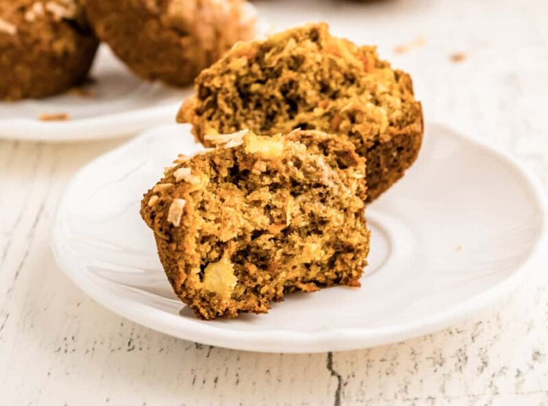 A morning glory muffin split in half sits on a white plate, revealing a moist, textured interior with visible chunks of fruit and nuts. In the background, more morning glory muffins rest slightly out of focus on another plate.