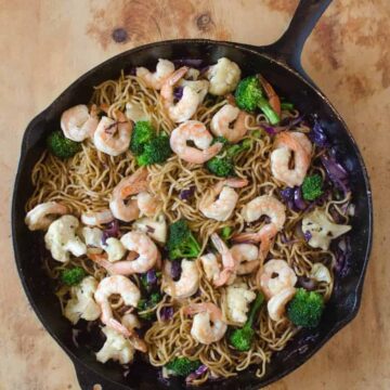 A cast iron skillet filled with a vibrant shrimp stir fry recipe—noodles, shrimp, broccoli florets, cauliflower, and purple cabbage—all mixed together and served on a wooden surface.