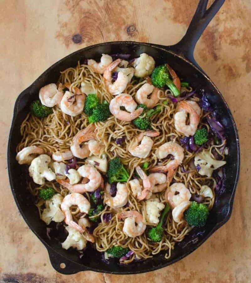 A cast iron skillet filled with a vibrant shrimp stir fry recipe—noodles, shrimp, broccoli florets, cauliflower, and purple cabbage—all mixed together and served on a wooden surface.