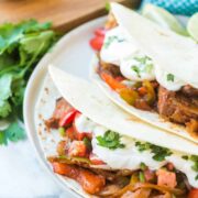 Two soft flour tortillas filled with sautéed onions, bell peppers, and seasoned steak fajitas recipe meat, topped with sour cream and fresh cilantro, served on a white plate with cilantro and lime wedges nearby.