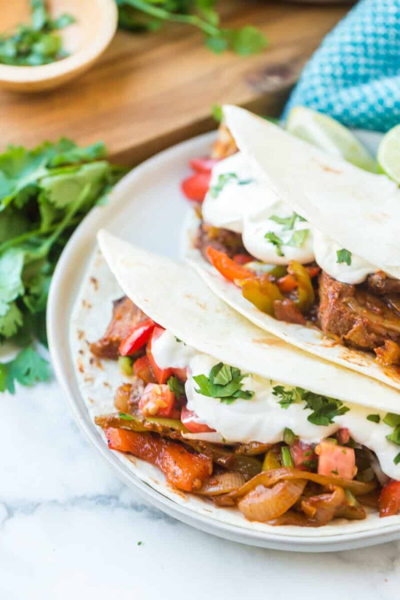 Two soft flour tortillas filled with sautéed onions, bell peppers, and seasoned steak fajitas recipe meat, topped with sour cream and fresh cilantro, served on a white plate with cilantro and lime wedges nearby.