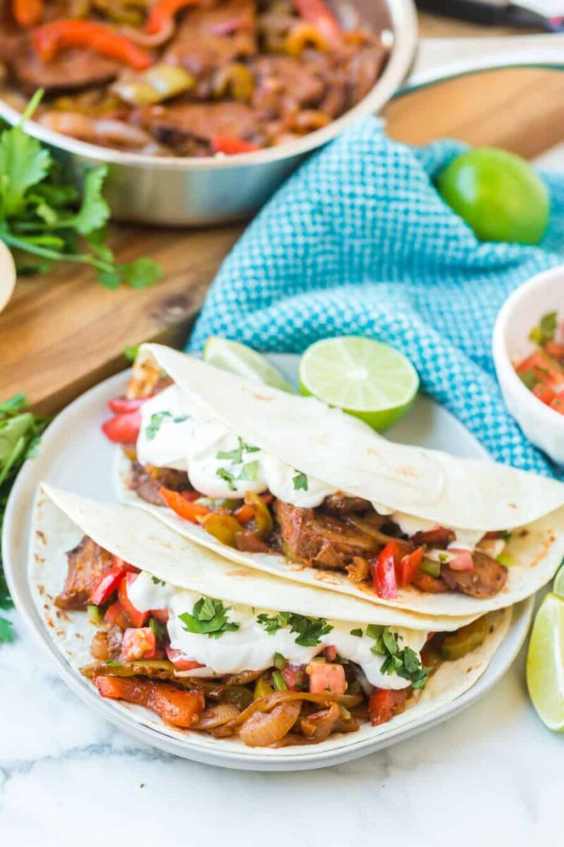 Two soft tortillas filled with sliced steak, sautéed bell peppers, onions, and diced tomatoes, topped with sour cream and cilantro—this steak fajitas recipe is served on a white plate with lime wedges. A skillet and herbs are in the background.