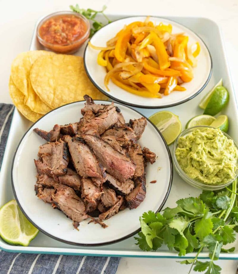 A plate with grilled steak slices, tortillas, guacamole, salsa, sautéed yellow bell peppers, lime wedges, and fresh cilantro on a tray—everything you need to try a delicious steak fajitas recipe at home.