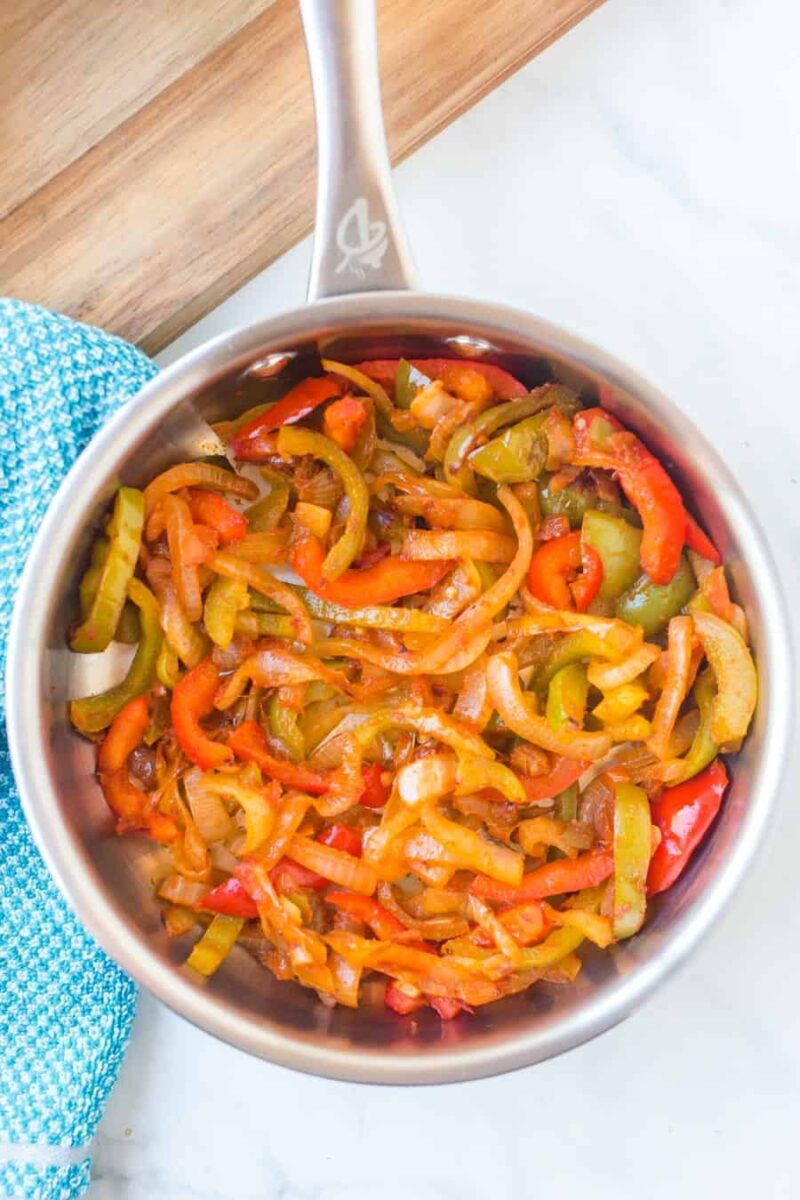 A stainless steel saucepan filled with cooked sliced onions, red and green bell peppers, and tomatoes—perfect ingredients for a steak fajitas recipe—rests on a marble countertop next to a blue textured cloth.