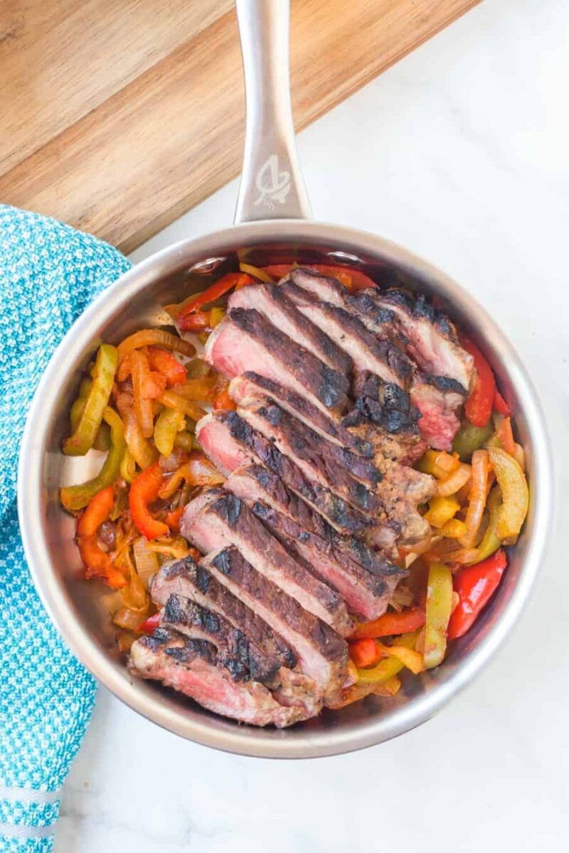 A stainless steel pan filled with sautéed bell peppers and onions, topped with sliced, grilled steak—perfect for your next steak fajitas recipe. The pan rests on a white surface beside a blue patterned towel and a wooden board.