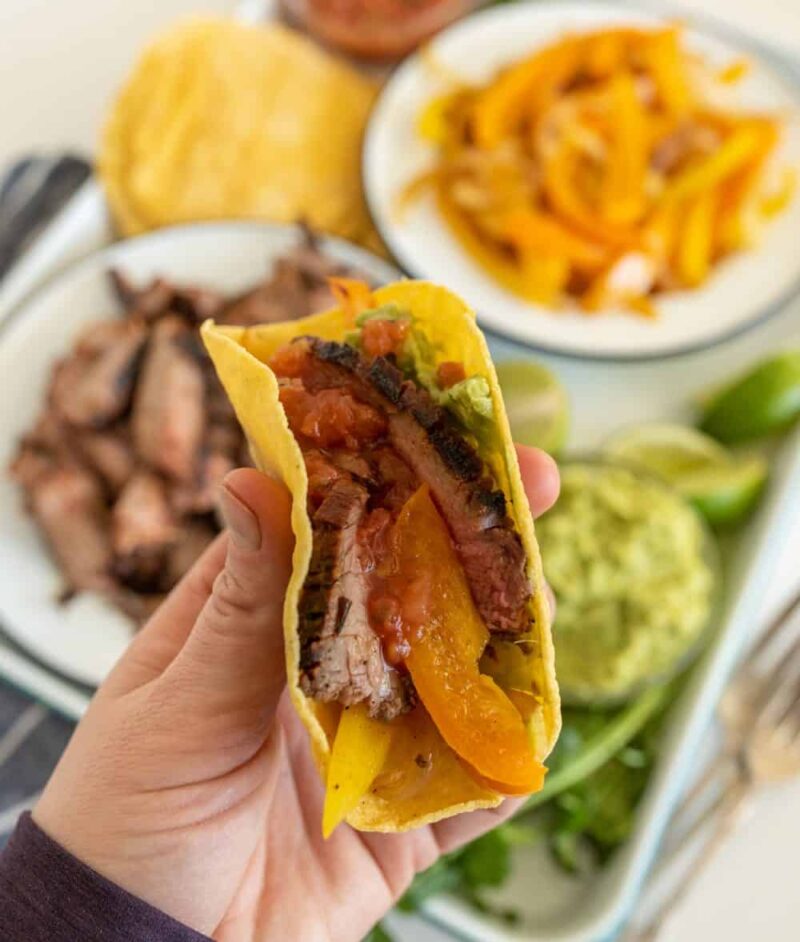 A hand holding a taco filled with grilled steak, yellow bell peppers, salsa, and guacamole—a delicious steak fajitas recipe—surrounded by plates of sliced steak, bell peppers, tortillas, guacamole, and lime in the background.