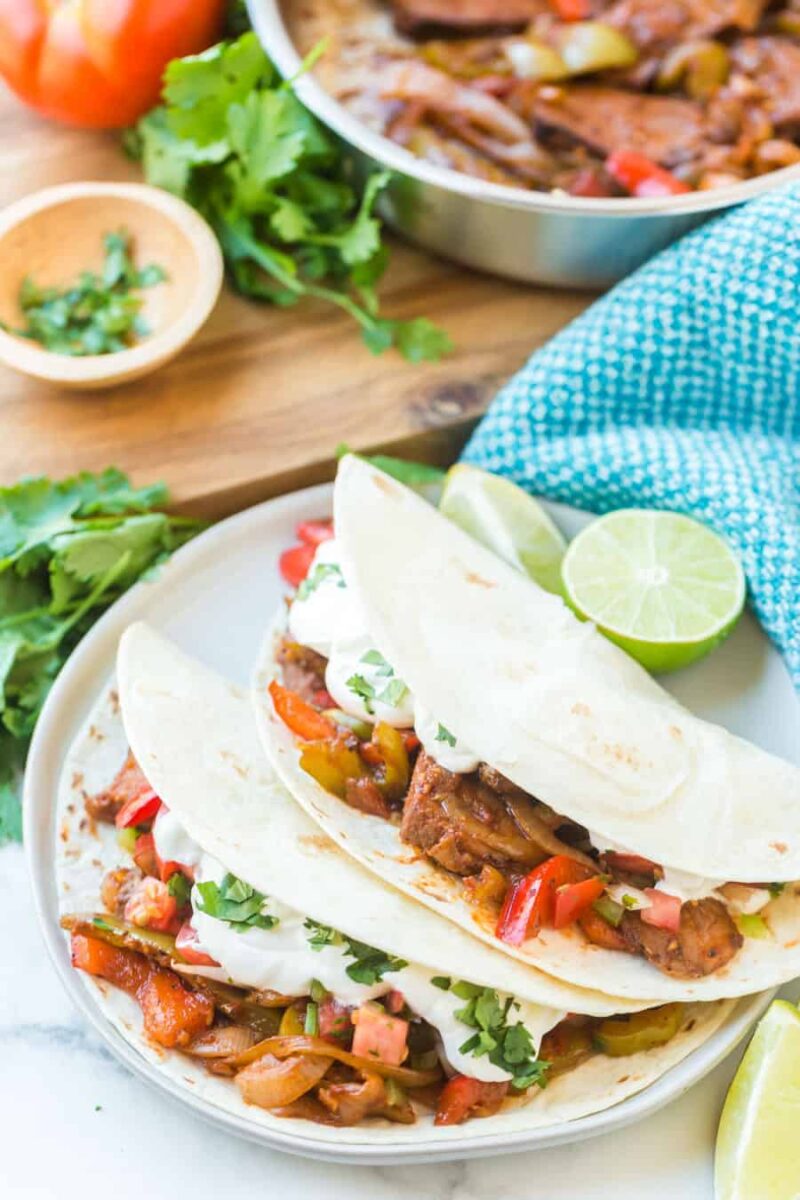 Three flour tortillas filled with seasoned grilled vegetables and tender steak, inspired by a classic steak fajitas recipe, topped with fresh cilantro and sour cream, served on a white plate with lime wedges. A skillet and fresh herbs are in the background.