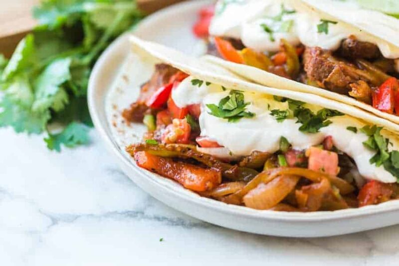 Two soft tacos filled with seasoned beef, sautéed onions, bell peppers, fresh salsa, and topped with sour cream and chopped cilantro—served on a white plate and inspired by a classic steak fajitas recipe.