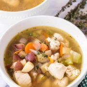 A white bowl filled with a comforting chicken stew recipe featuring chunks of chicken, potatoes, carrots, celery, and herbs. Another bowl and thyme sprigs sit in the background beside a green checkered cloth.