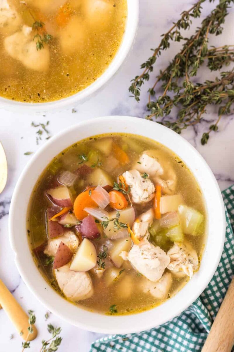 Two bowls of homemade chicken vegetable soup, inspired by a classic chicken stew recipe, with chunks of chicken, carrots, potatoes, celery, and herbs. Fresh thyme and a green gingham napkin sit beside the bowls on a white marble surface.