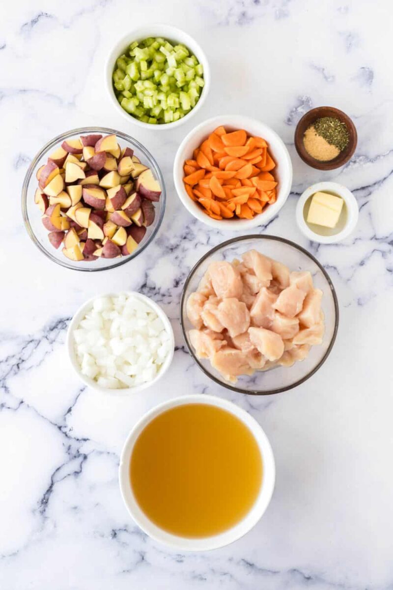 Bowls of chopped ingredients for a chicken stew recipe on a marble surface: diced potatoes, celery, carrots, onions, raw chicken pieces, butter, seasonings, and a bowl of broth.