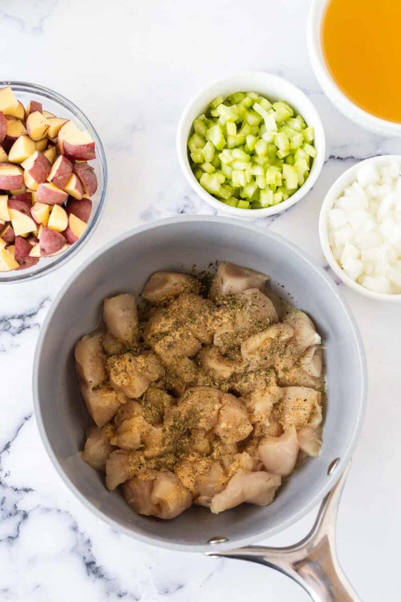 A saucepan filled with diced raw chicken and seasonings is surrounded by bowls of chopped red potatoes, celery, and onions on a white marble surface—perfect ingredients for a hearty chicken stew recipe. A bowl of broth sits in the top right corner.