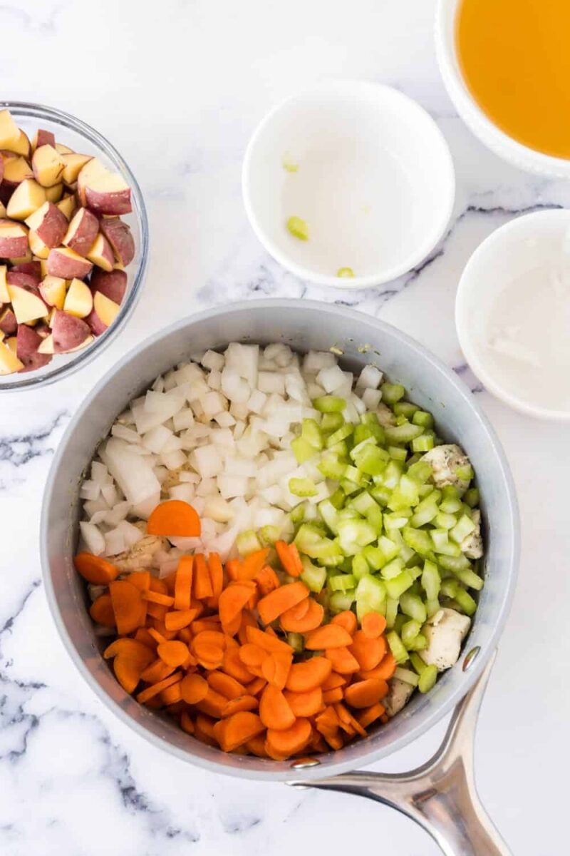 A saucepan with chopped onions, celery, and carrots sits next to bowls of diced red potatoes and broth—perfect ingredients for a comforting chicken stew recipe—all arranged on a white marble surface.