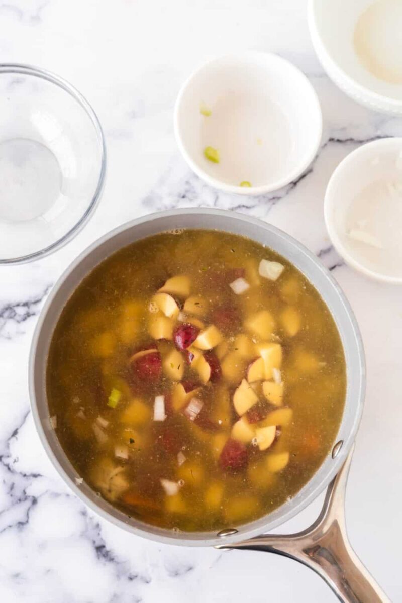 A saucepan filled with a hearty chicken stew recipe—brimming with chopped potatoes, onions, and broth—sits on a marble countertop. Empty bowls are nearby, some with food remnants.