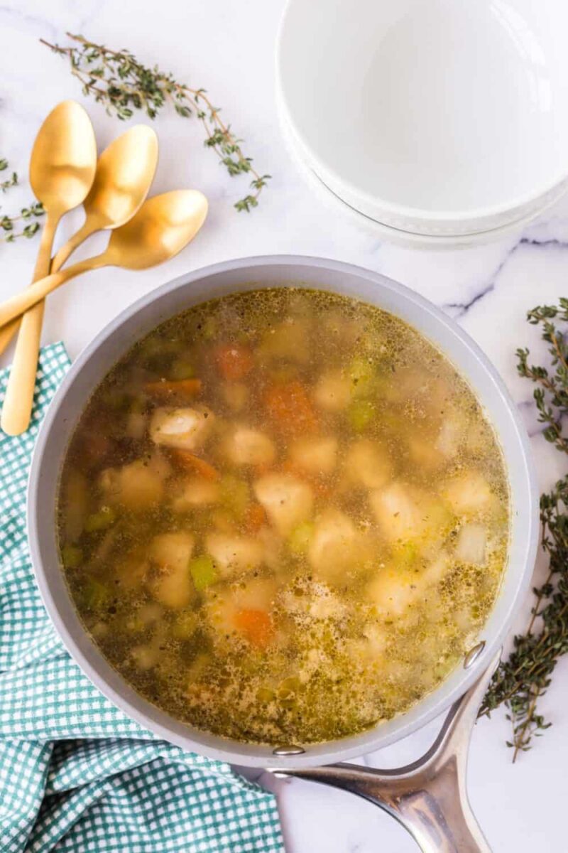 A pot of hearty vegetable soup, reminiscent of a classic chicken stew recipe, with carrots, celery, and potatoes in broth sits on marble. Gold spoons, a green checkered cloth, fresh thyme sprigs, and stacked white bowls complete the scene.