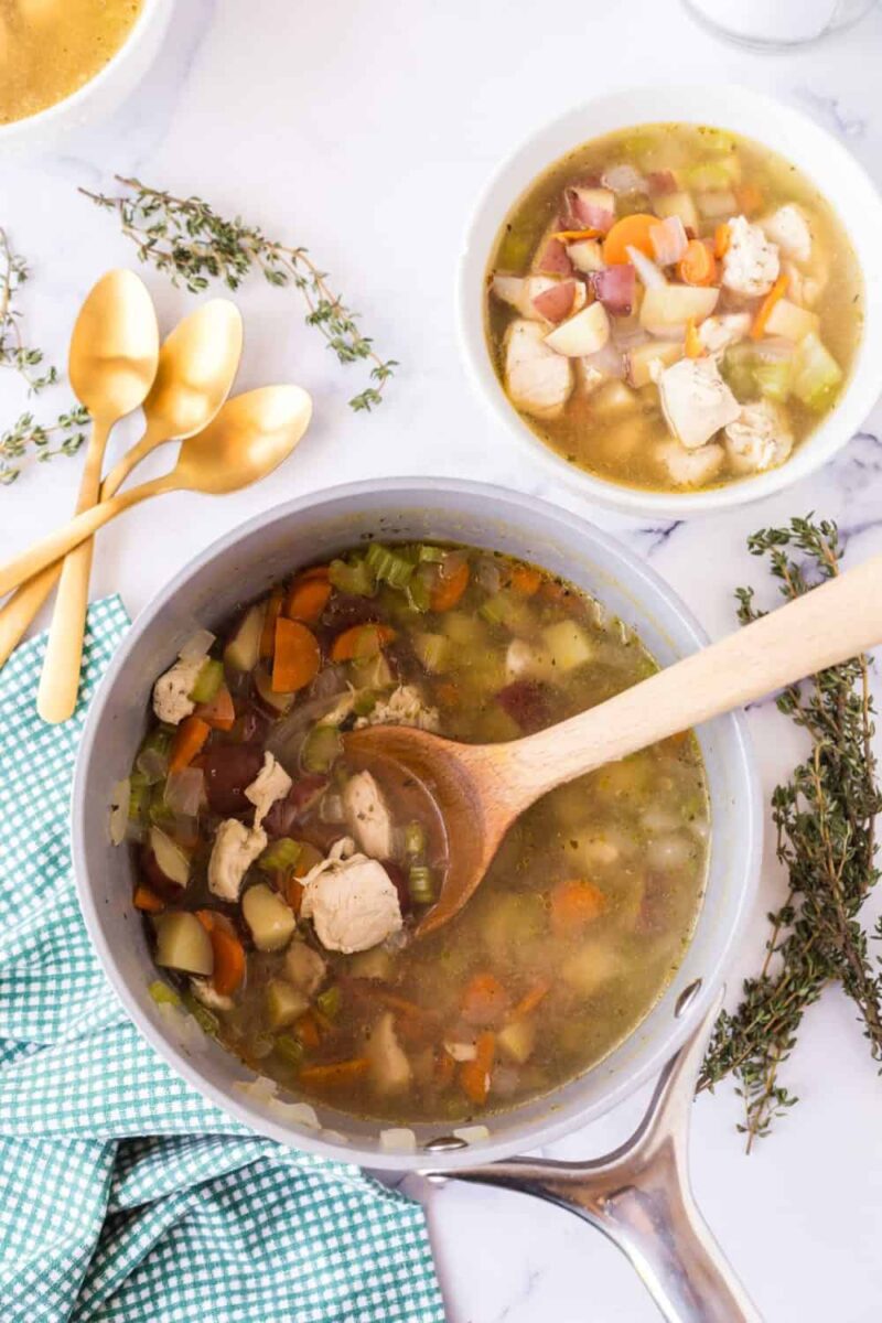 A pot of hearty chicken stew recipe with chunks of chicken, carrots, potatoes, and celery sits on a white surface. A wooden spoon rests inside, with a bowl of the soup, three gold spoons, and sprigs of thyme nearby.