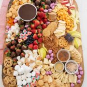 A large wooden chocolate charcuterie board filled with assorted cheeses, crackers, cookies, chocolate-covered strawberries, apple slices, grapes, marshmallows, pretzels, and three small bowls of dips or sauces.