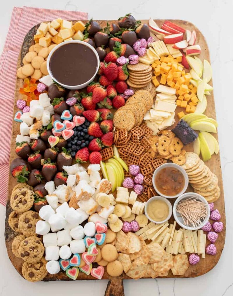 A large wooden chocolate charcuterie board filled with assorted cheeses, crackers, cookies, chocolate-covered strawberries, apple slices, grapes, marshmallows, pretzels, and three small bowls of dips or sauces.