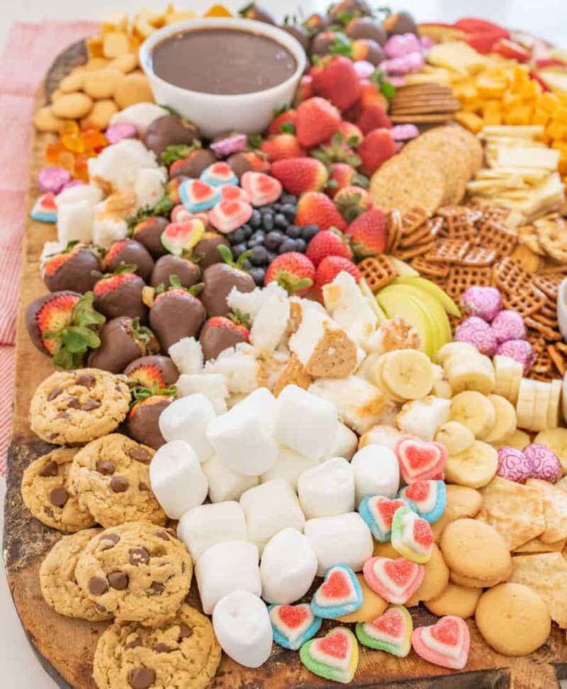 A large chocolate charcuterie board filled with assorted treats, including chocolate chip cookies, marshmallows, heart-shaped gummies, chocolate-covered strawberries, fresh fruits, pretzels, crackers, cookies, and a bowl of chocolate dip.