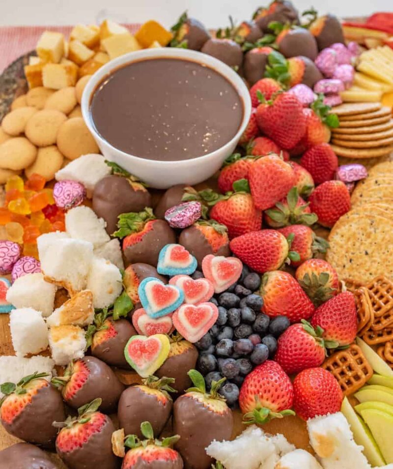 A chocolate charcuterie board featuring a bowl of chocolate fondue, surrounded by strawberries, blueberries, chocolate-covered strawberries, heart-shaped gummies, cookies, pretzels, cheeses, and pink-wrapped candies.