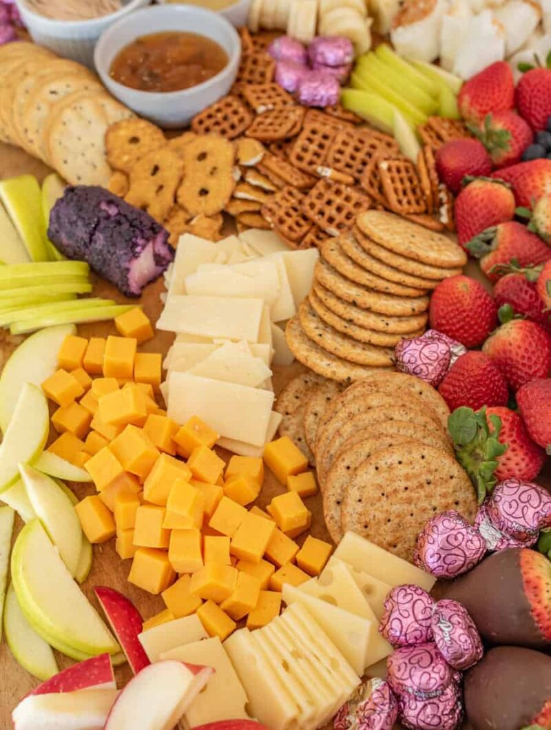 A chocolate charcuterie board featuring assorted cheeses, crackers, pretzels, apple slices, celery, strawberries, chocolate-covered treats, and bowls of dip—all artfully arranged on a wooden surface.