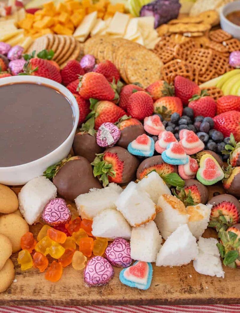 A colorful chocolate charcuterie board with strawberries, blueberries, cheese, crackers, cookies, chocolate-dipped strawberries, gummy candies, heart-shaped marshmallows, chocolate eggs, coconut squares, and a bowl of chocolate dip.