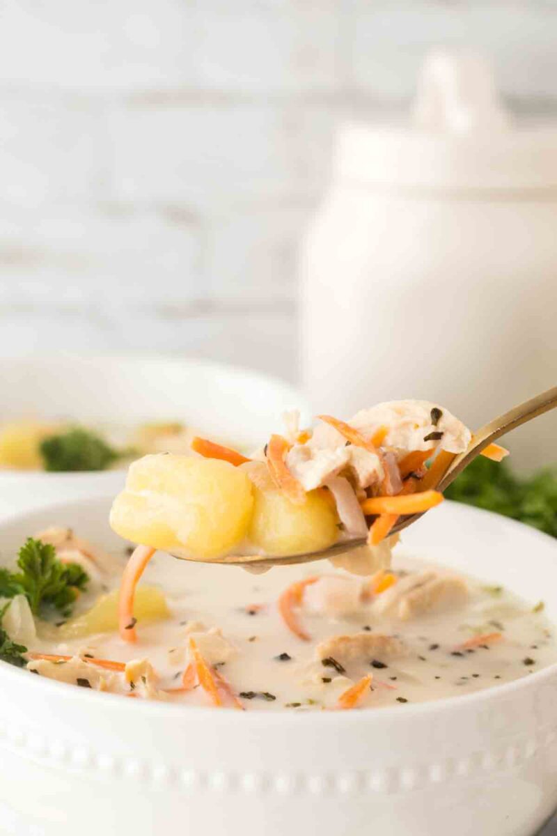 A close-up of a spoonful of creamy Italian chicken gnocchi soup with potatoes, shredded carrots, and herbs, held above a white bowl filled with the same soup. A sprig of parsley sits next to the bowl.