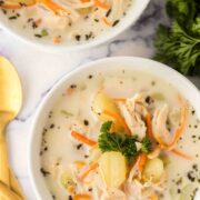 Two white bowls filled with creamy Italian chicken gnocchi soup, featuring shredded chicken, gnocchi, carrot strips, and fresh parsley. A gold spoon and fork rest beside the bowls on a white surface.