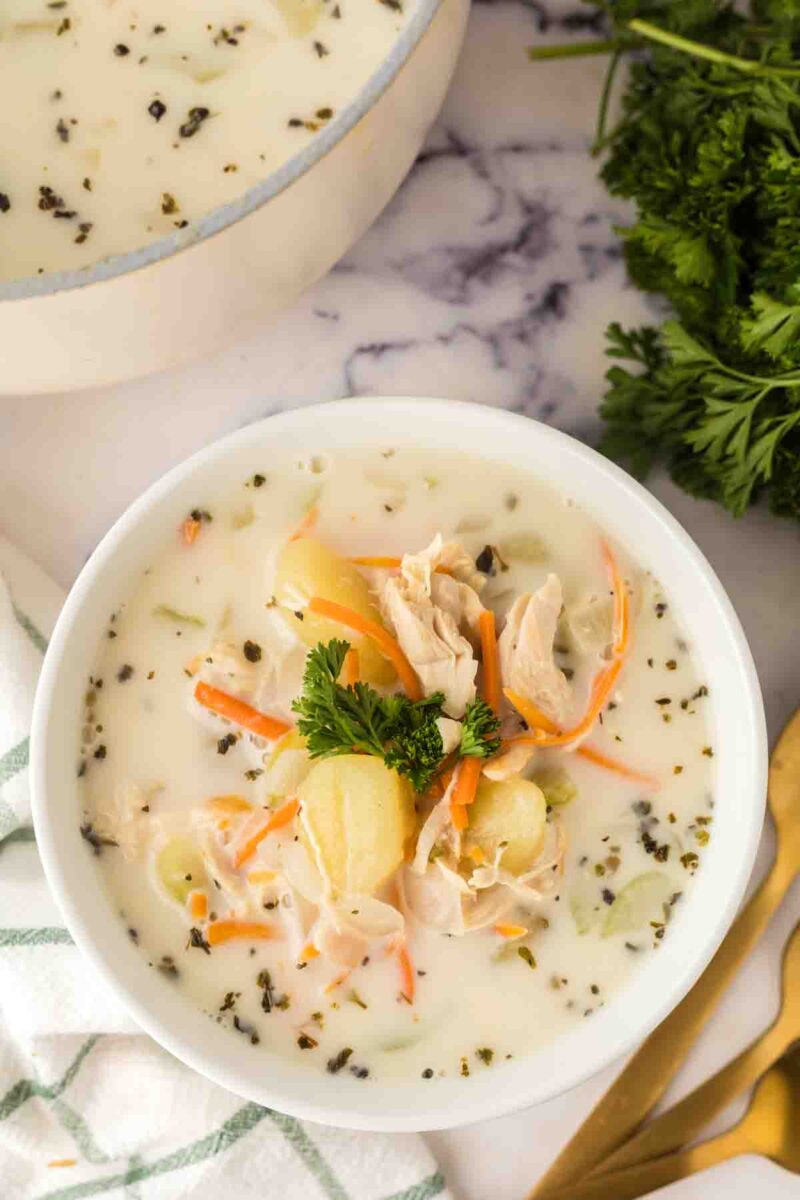 A bowl of creamy Italian chicken gnocchi soup with potatoes, carrots, celery, herbs, and shredded chicken, garnished with fresh parsley, sits on a marble surface beside a white pot, parsley bunch, and gold utensils.