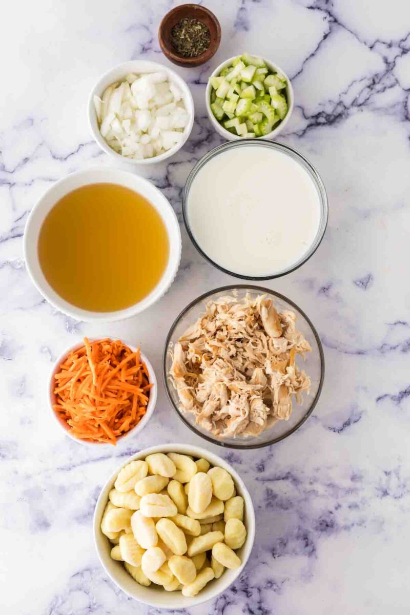 A top-down view of separated ingredients for Italian chicken gnocchi soup on a marble surface, including shredded chicken, gnocchi, carrots, onions, celery, broth, cream, and Italian herbs in small bowls.