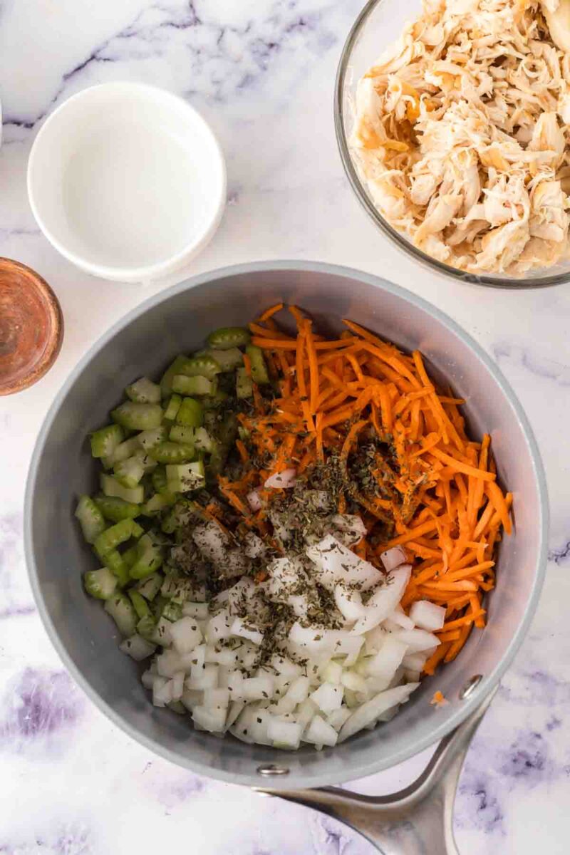 A saucepan on a marble surface contains chopped celery, shredded carrots, diced onions, and herbs—the perfect base for Italian chicken gnocchi soup. Nearby are bowls of shredded chicken and a small dish of water.