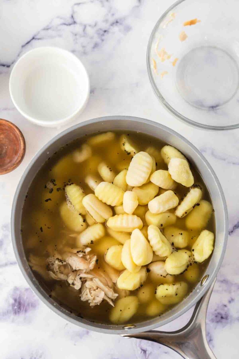 A saucepan filled with italian chicken gnocchi soup—brimming with chicken broth, shredded chicken, and pillowy gnocchi—sits on a marble countertop beside an empty glass bowl and a small white dish.
