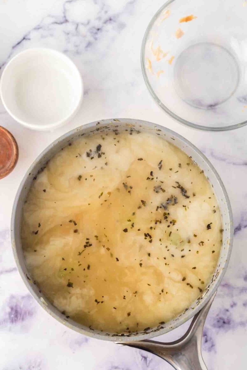 A saucepan filled with Italian chicken gnocchi soup, its light brothy base and herbs visible, sits on a marble countertop. Nearby are an empty glass bowl, a small white bowl, and a copper-colored container.