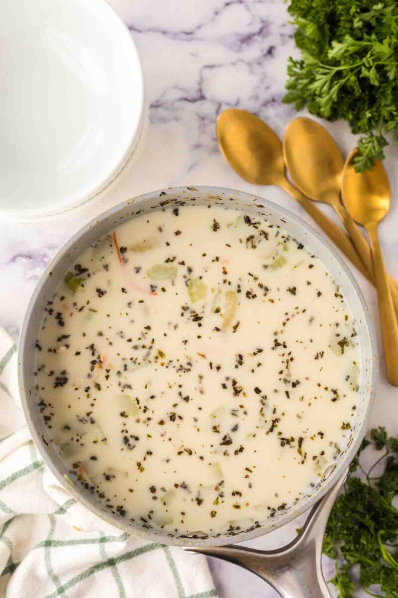 A pot of creamy Italian chicken gnocchi soup with visible herbs and vegetables sits on a marble countertop near a white bowl, three gold spoons, a green-striped towel, and fresh parsley.