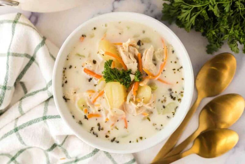 A bowl of creamy Italian chicken gnocchi soup with shredded chicken, gnocchi, carrots, and herbs, garnished with parsley. Gold spoons and a green-checked napkin sit beside the bowl on a marble surface.