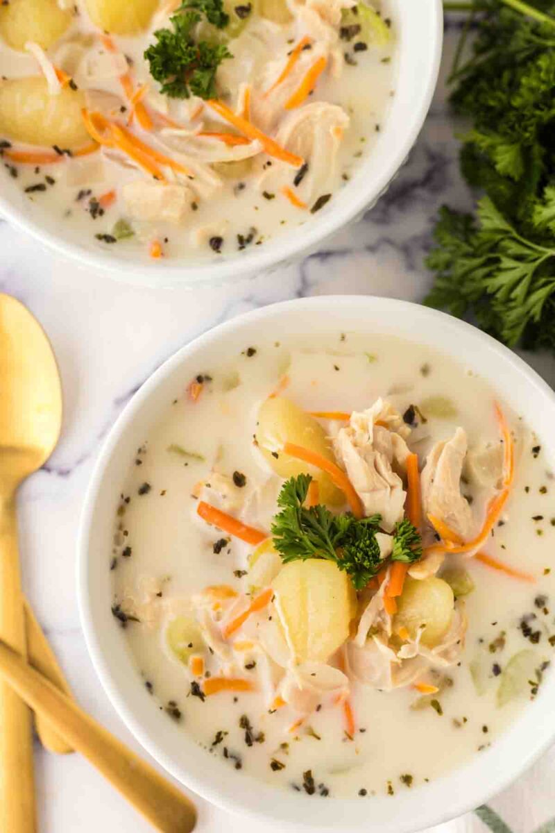 Two white bowls filled with creamy Italian chicken gnocchi soup, featuring shredded chicken, gnocchi, carrot strips, and fresh parsley. A gold spoon and fork rest beside the bowls on a white surface.