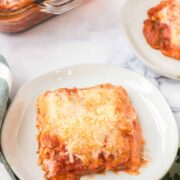 A serving of cheesy baked lasagna with spinach and ricotta, topped with tomato sauce and melted cheese on a white plate. A baking dish of lasagna and another plate sit in the background, with fresh parsley and a striped towel nearby.