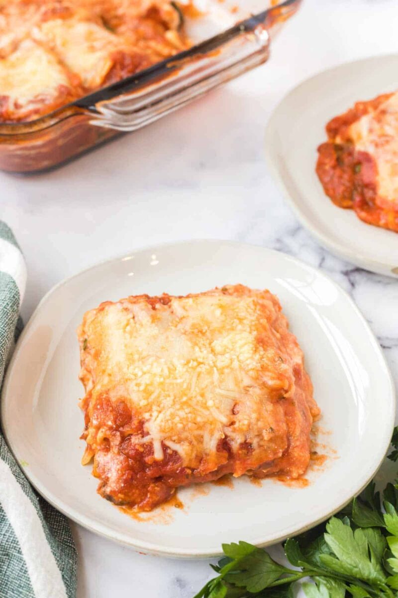 A serving of cheesy baked lasagna with spinach and ricotta, topped with tomato sauce and melted cheese on a white plate. A baking dish of lasagna and another plate sit in the background, with fresh parsley and a striped towel nearby.