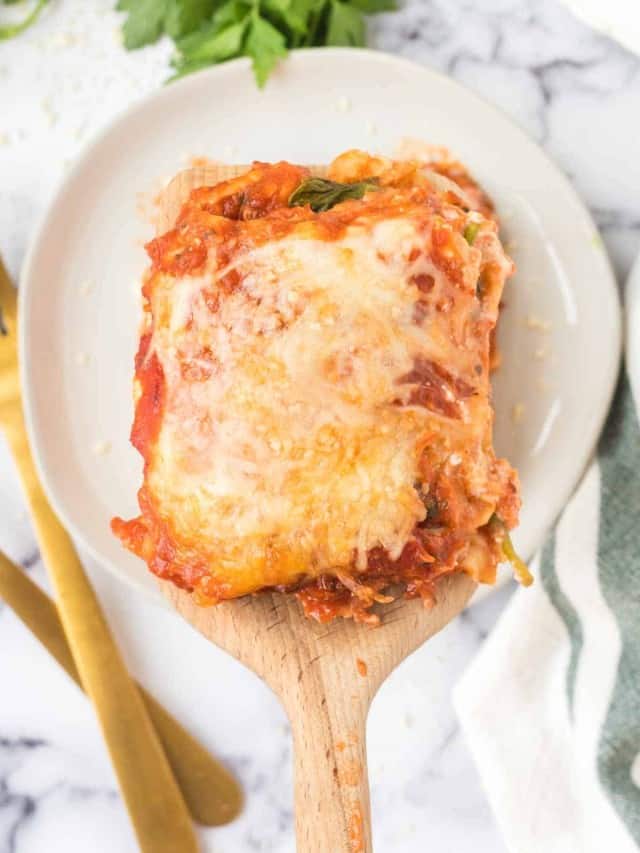 A spatula holds a cheesy slice of baked lasagna with spinach and ricotta over a white plate. The lasagna is topped with melted cheese and tomato sauce. Gold utensils, a green herb, and a white napkin are visible nearby.