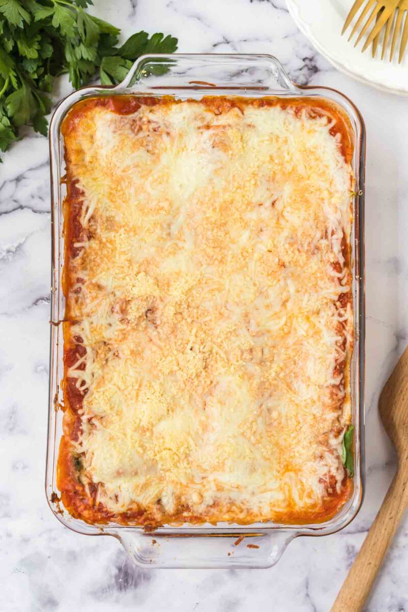 A glass baking dish filled with lasagna with spinach and ricotta, topped with melted, golden cheese, sits on a marble surface next to a wooden spoon, a white plate, and fresh parsley.
