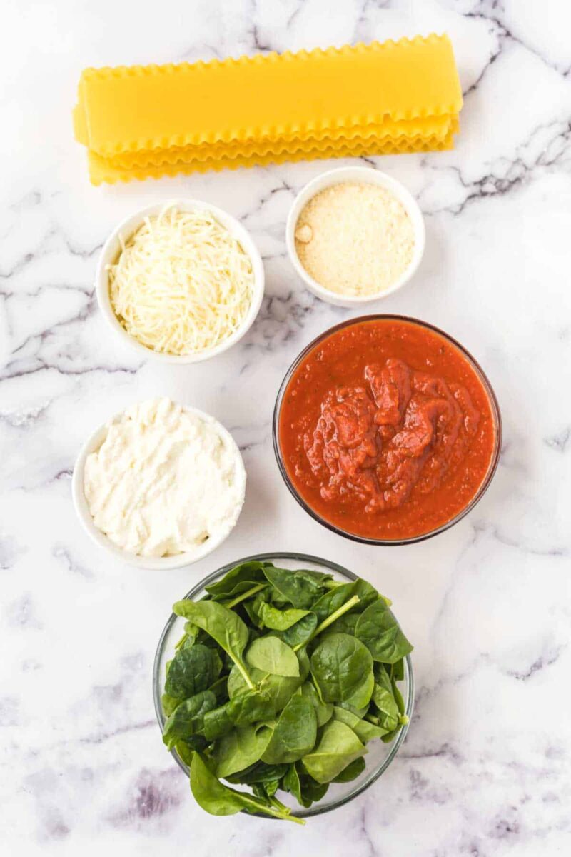 Overhead view of lasagna with spinach and ricotta: noodles, shredded mozzarella, grated parmesan, ricotta cheese, tomato sauce, and fresh spinach arranged separately on a white marble surface.
