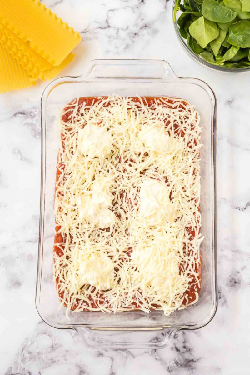 A glass baking dish with lasagna layers topped with tomato sauce, shredded cheese, and dollops of ricotta. Uncooked lasagna noodles and a bowl of fresh spinach hint at a lasagna with spinach and ricotta on the marble countertop nearby.