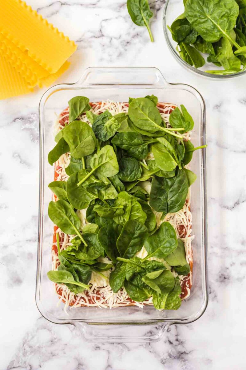 A glass baking dish with layers of lasagna noodles, shredded cheese, red sauce, and fresh spinach leaves—perfect for making lasagna with spinach and ricotta. Nearby are dry noodles and a bowl of fresh spinach on a marble countertop.